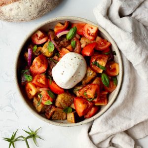 Burrata panzanella salad in a bowl surrounded by loaf of bread, grey dish towel, and a tomato