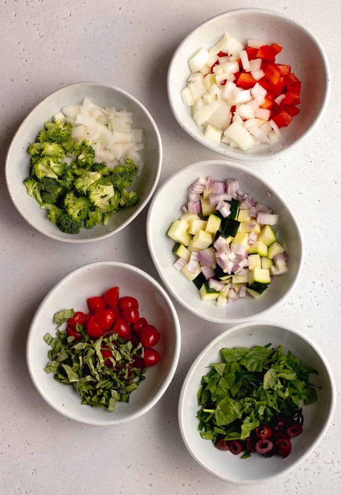 Five small bowls with different raw vegetables on a grey backdrop