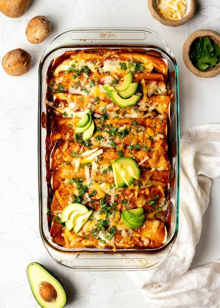 Glass baking dish with mushroom enchiladas topped with avocado slices, with ingredients surrounding the tray on a white backdrop