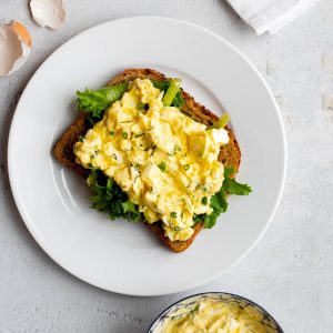 healthy egg salad on toast with lettuce on a plate. small bowl of leftover egg salad is to the side