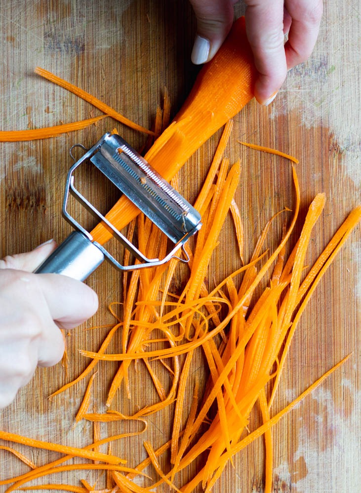 hands using a julienne peeler to julienne a carrot on wooden cutting board