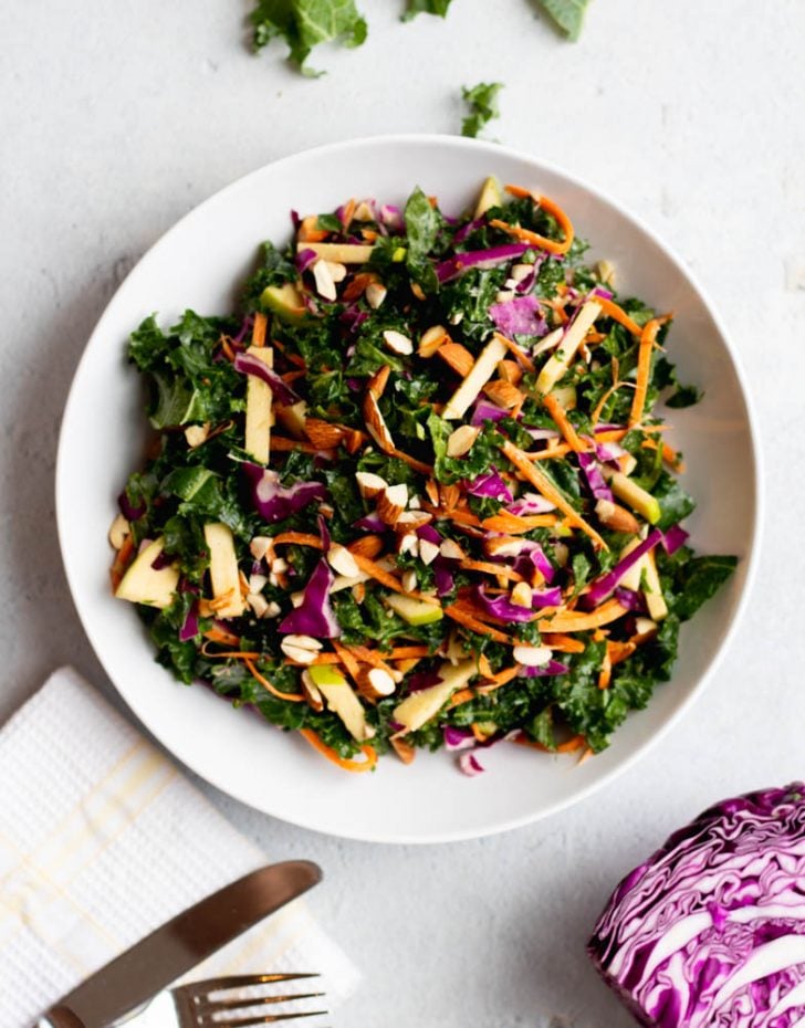 white bowl filled with kale slaw salad, surrounded by a few ingredients and a white napkin with a fork and knife