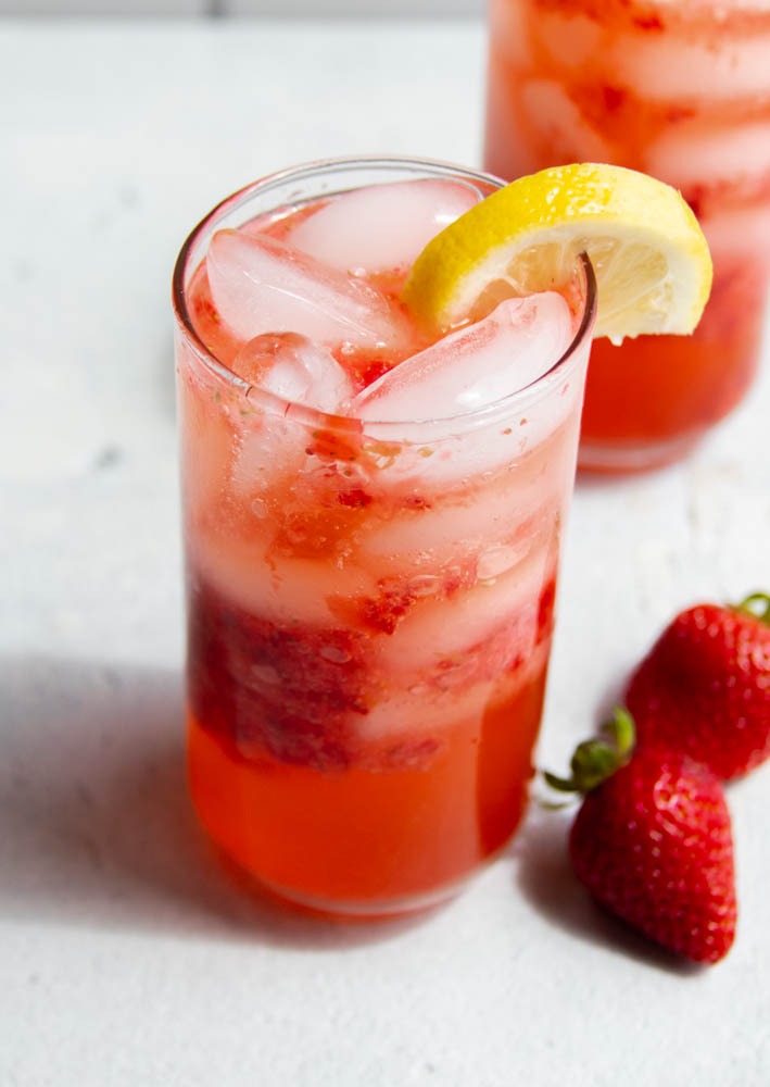 two glasses of strawberry lemon mocktail on a light grey surface with two strawberries 