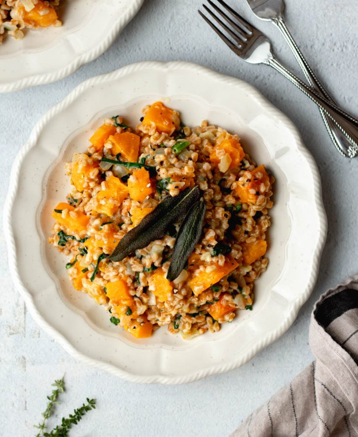 White plate with butternut squash farro risotto garnished with fried sage leaves on a light grey backdrop
