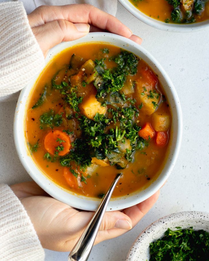 hands surrounding a bowl of carrot potato soup on a grey backdrop