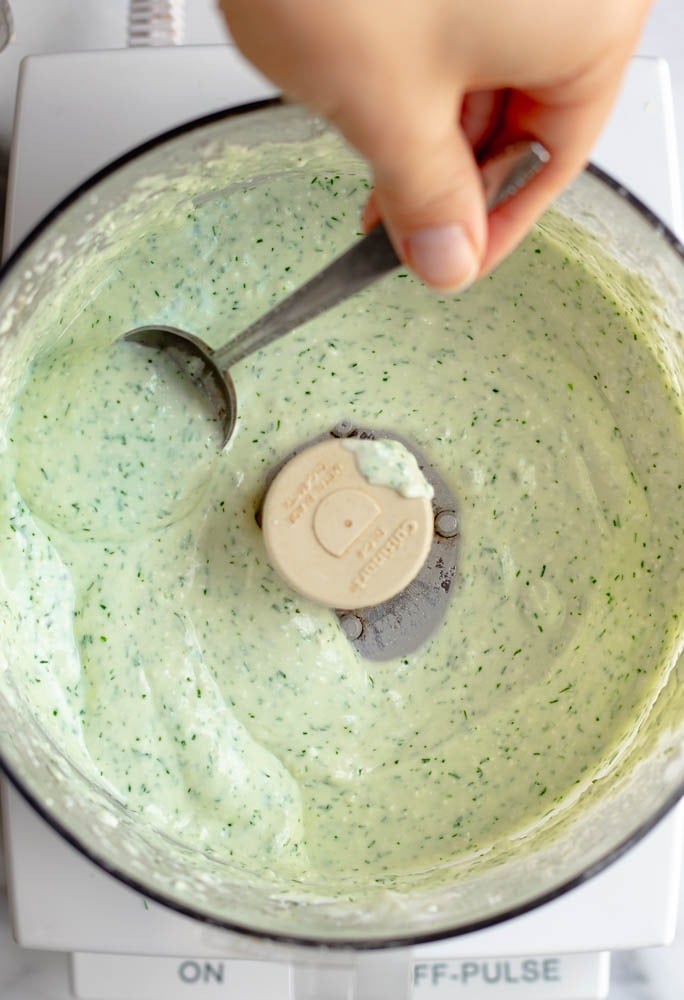 hand scooping up some feta dill dressing with a spoon from a food processor