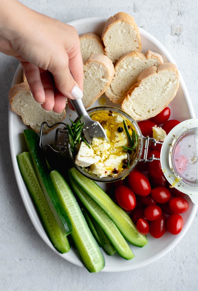 Hand scooping out marinated feta from a jar with a cheese knife, that is arranged on a serving plate with vegetables and bread
