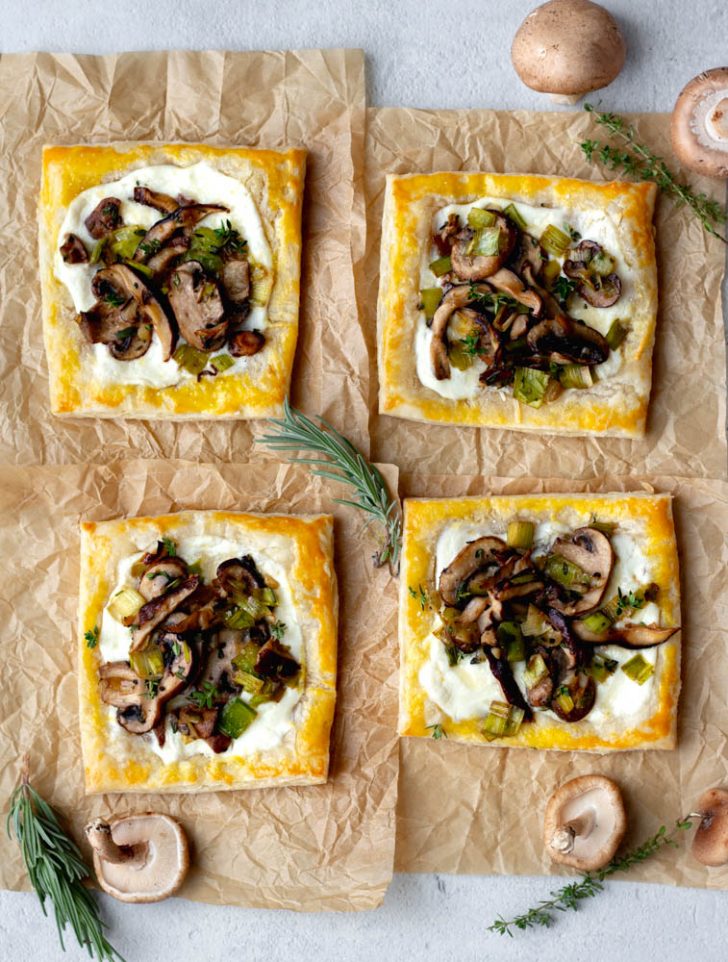 four mushroom puff pastry tarts on brown parchment paper with sprigs of fresh herbs
