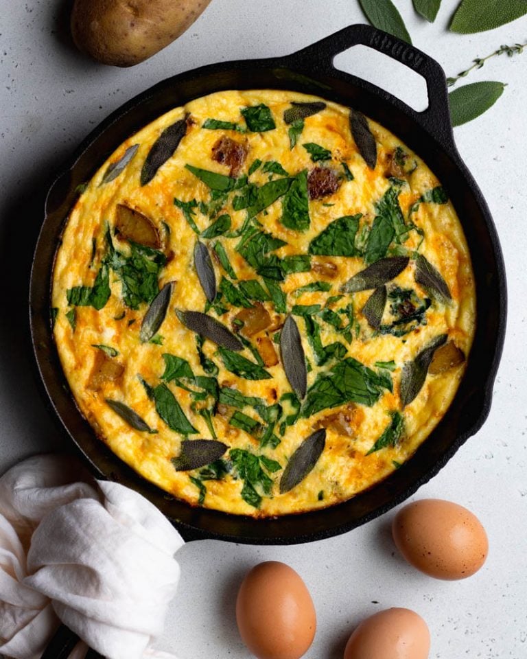 spinach potato frittata in a cast iron skillet on a light grey backdrop surrounded by a few ingredients
