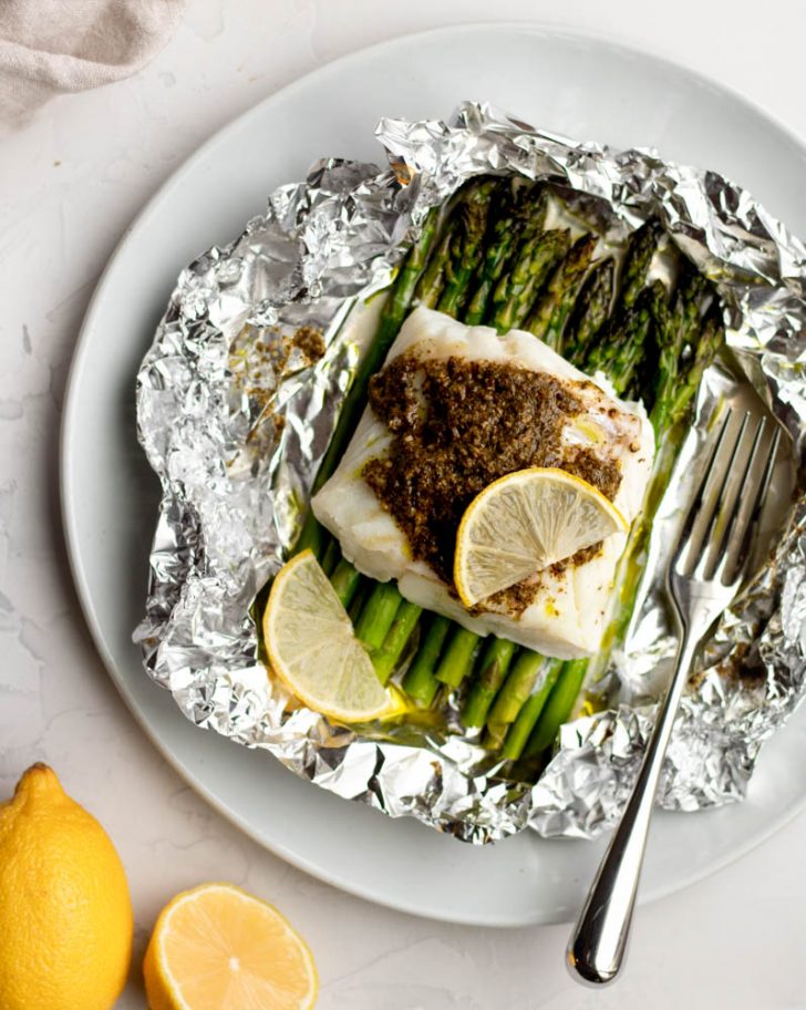 piece of cod fish with pesto and a lemon slice over a bed of asparagus in an opened foil packet