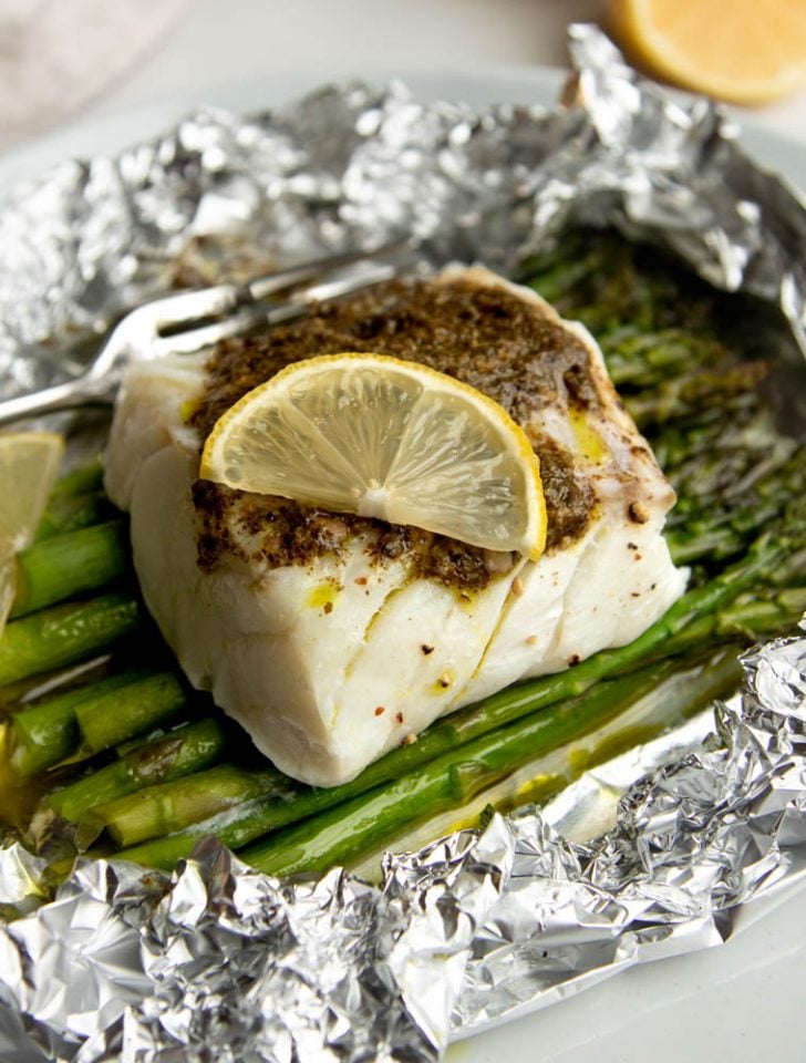 piece of cod fish with pesto and a lemon slice over a bed of asparagus in an opened foil packet