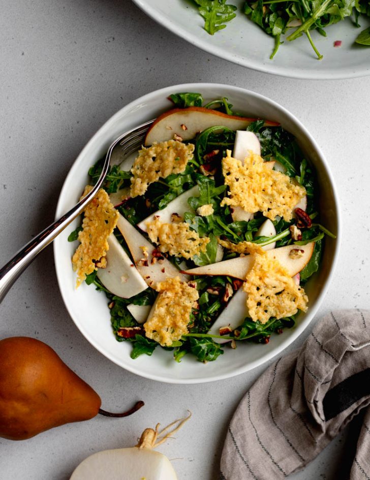 green salad with parmesan crisps in a white bowl with a fork, next to a dish towel, pear, and turnip