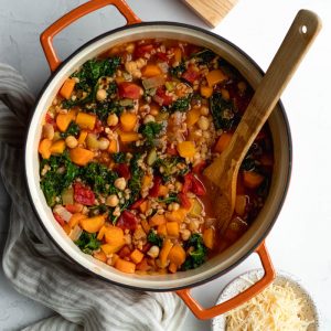 Large pot of farro vegetable soup with a wooden mixing spoon next to a small bowl of parmesan cheese