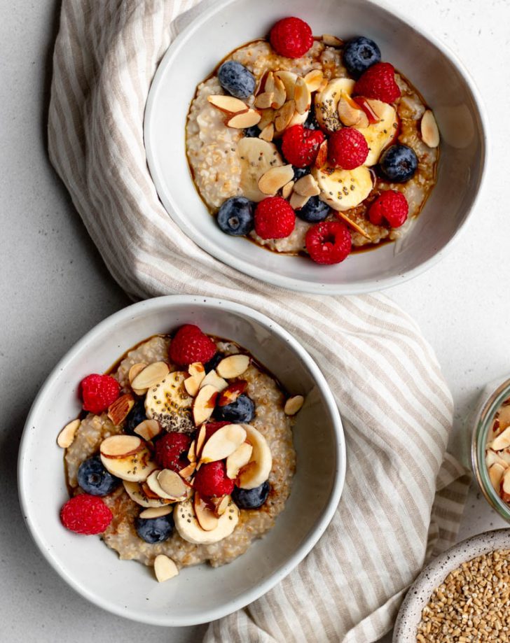 Two bowls of steel cut oatmeal with berries, bananas, and almonds, on a light grey backdrop with a striped dish towel