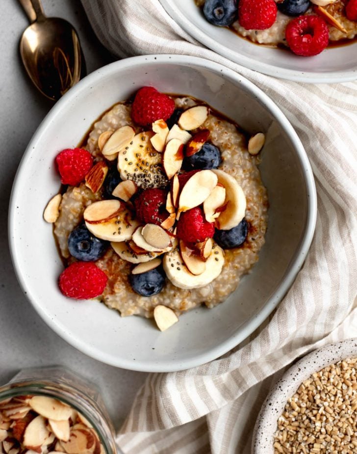 bowl of steel cut oatmeal topped with sliced almonds and berries, surrounded by small bowls of ingredients, a striped dish towel, and a spoon