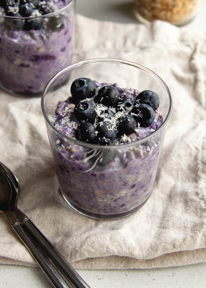 Glass cup filled with blueberry pie overnight oats garnished with fresh blueberries on a light beige dish towel