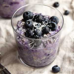 Glass cup filled with blueberry pie overnight oats garnished with fresh blueberries on a light beige dish towel