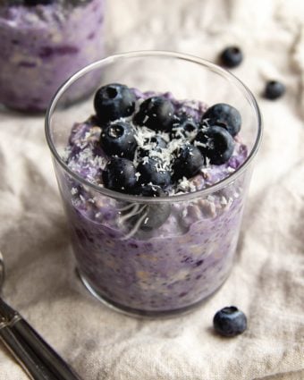 Glass cup filled with blueberry pie overnight oats garnished with fresh blueberries on a light beige dish towel