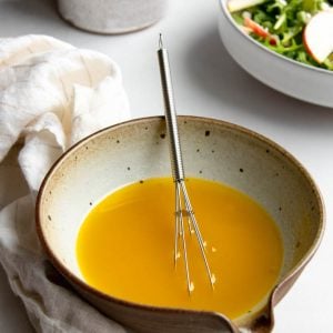 small bowl of vinaigrette on a light grey backdrop with a salad and olive oil decanter in the background