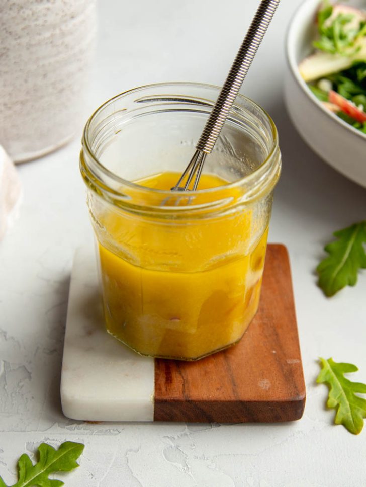 small jar of champagne vinaigrette on a marble and wooden coaster surrounded by arugula leaves