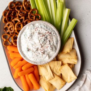 platter of veggies, pita chips, and pretzels with a bowl of yogurt dip in the center. There is a small side plate and ingredients next to the platter