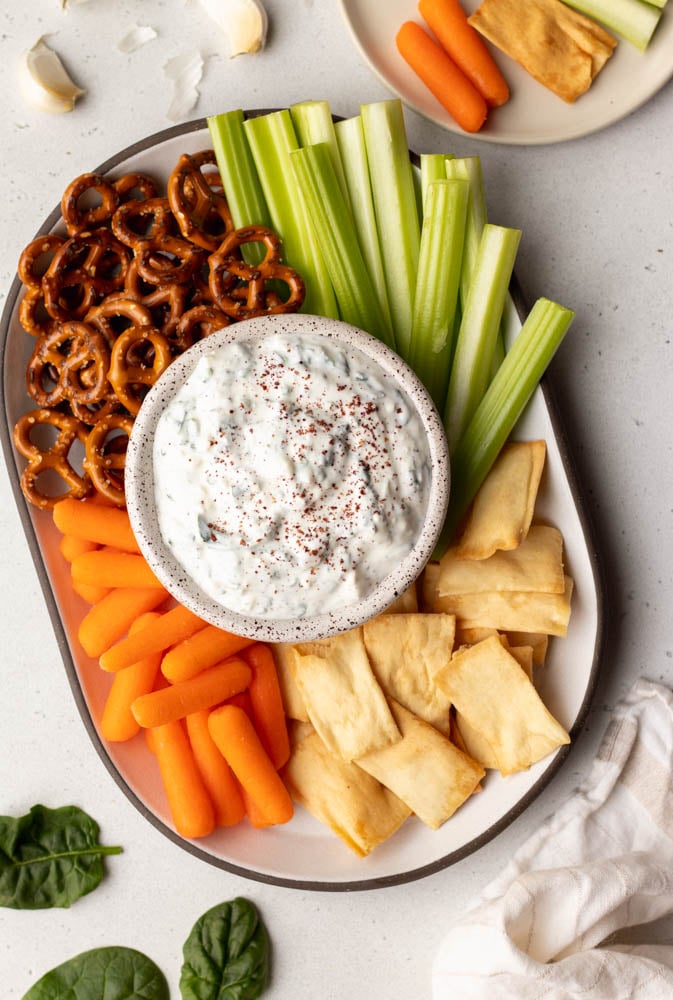 platter of veggies, pita chips, and pretzels with a bowl of yogurt dip in the center. There is a small side plate and ingredients next to the platter