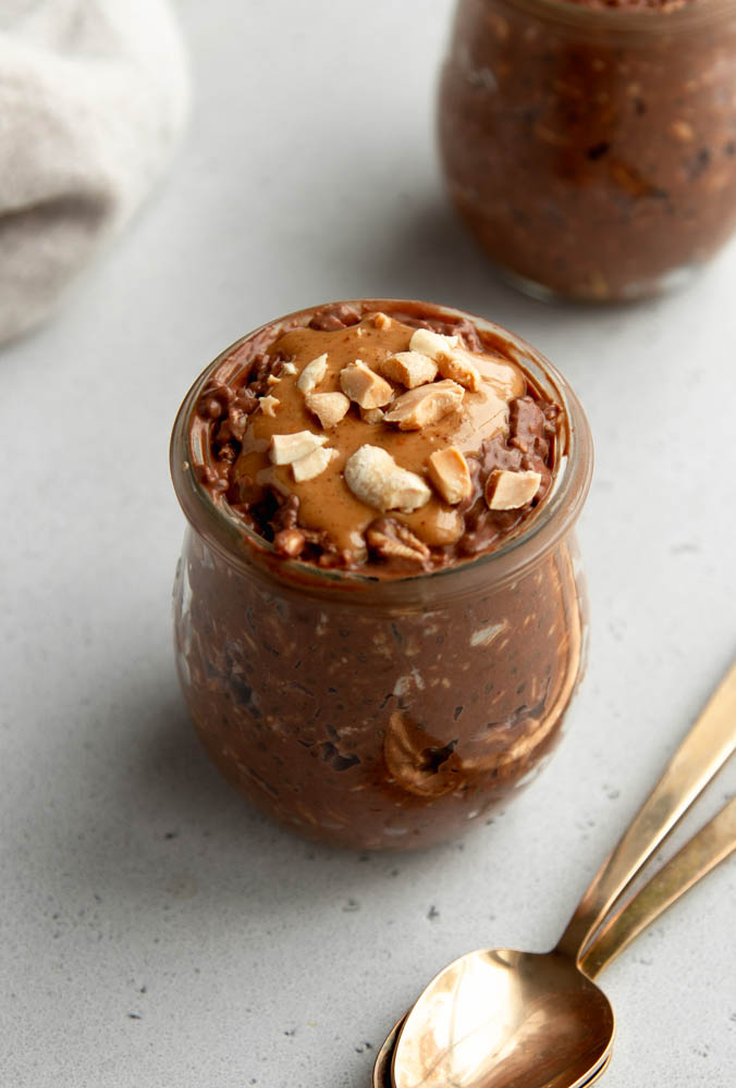 jar of chocolate peanut butter overnight oats with two gold spoons on a light grey backdrop