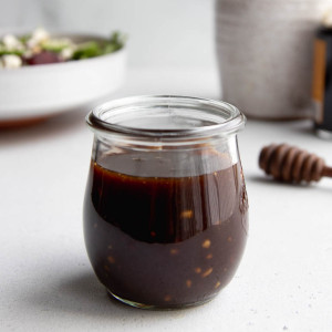 small jar of honey balsamic vinaigrette on a white surface with a salad and bottles of oil and vinegar in the background