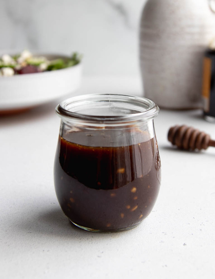 small jar of honey balsamic vinaigrette on a white surface with a salad and bottles of oil and vinegar in the background
