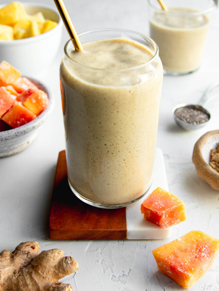 Pineapple papaya smoothie on a wooden and marble coaster with ingredients surrounding the cup