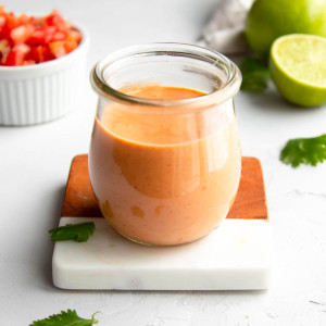 small jar of taco salad dressing on a white marble and wooden coaster with ingredients in the background