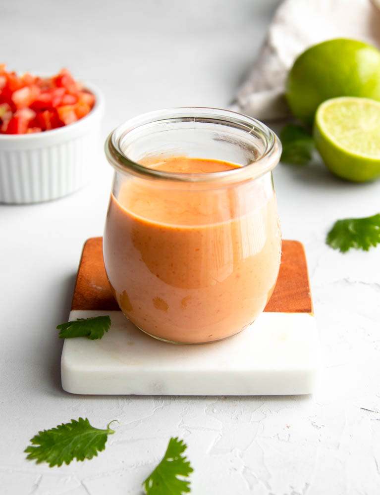 small jar of taco salad dressing on a white marble and wooden coaster with ingredients in the background