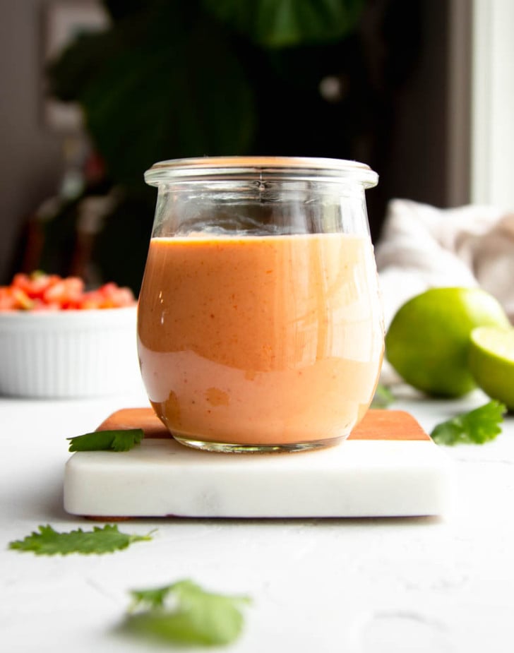 small jar of taco salad dressing on a white marble and wooden coaster with ingredients in the background