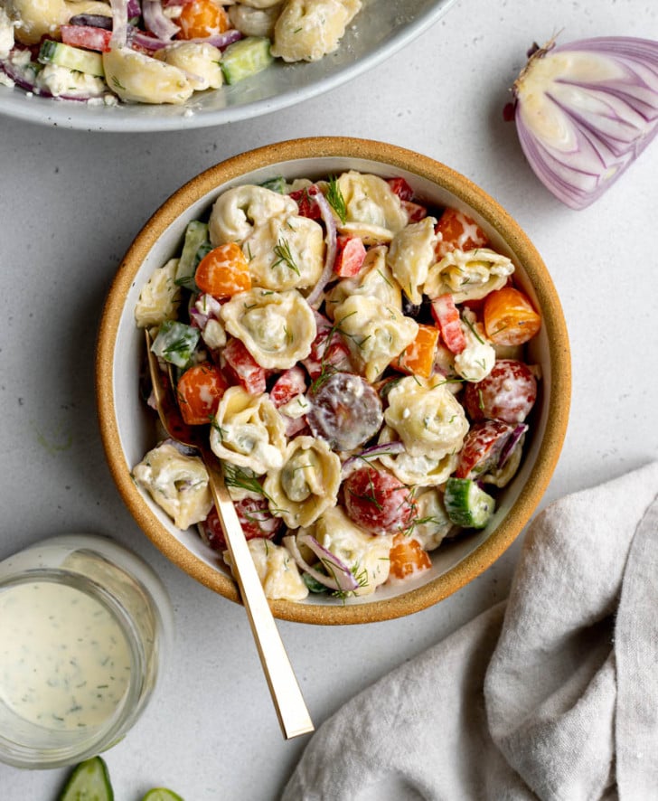 bowl of greek tortellini salad on a light grey backdrop surrounded by ingredients 