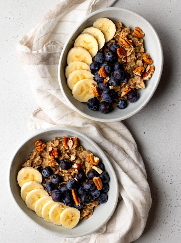 two bowls of oatmeal topped with banana, blueberries, and nuts on a light grey backdrop