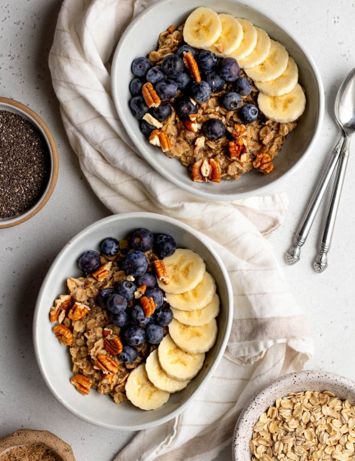 two bowls of oatmeal topped with banana, blueberries, and nuts on a light grey backdrop, with bowls of ingredients surrounding