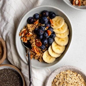 bowl of oatmeal topped with banana, blueberries, and nuts on a light grey backdrop, with bowls of ingredients surrounding
