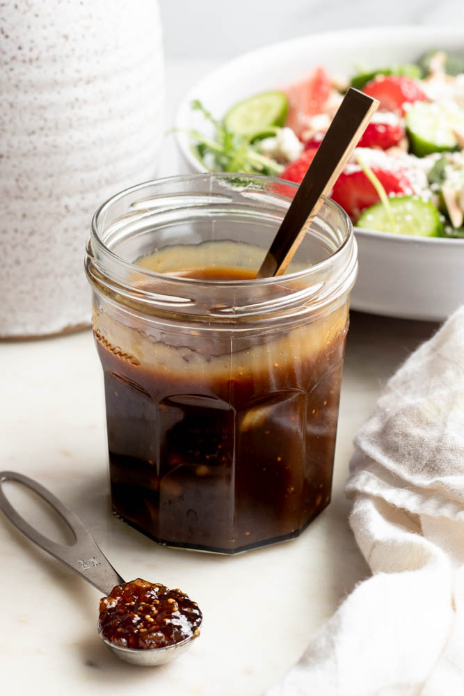 Jar of fig balsamic dressing with a spoon, with a salad and olive oil bottle in the background
