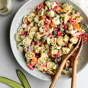 Greek tortellini salad mixed together in a large mixing bowl with wooden salad spoons surrounded by some ingredients