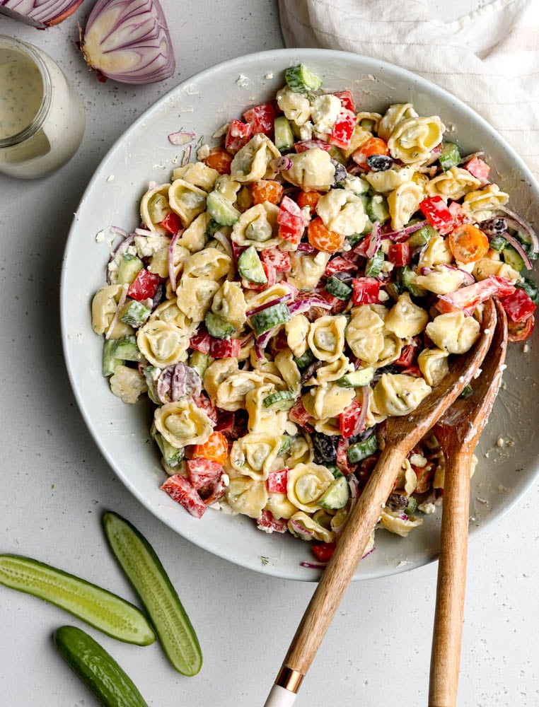 Greek tortellini salad mixed together in a large mixing bowl with wooden salad spoons surrounded by some ingredients