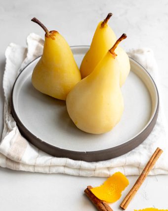 three poached pears on a white plate with cinnamon stick and orange peel at the foreground