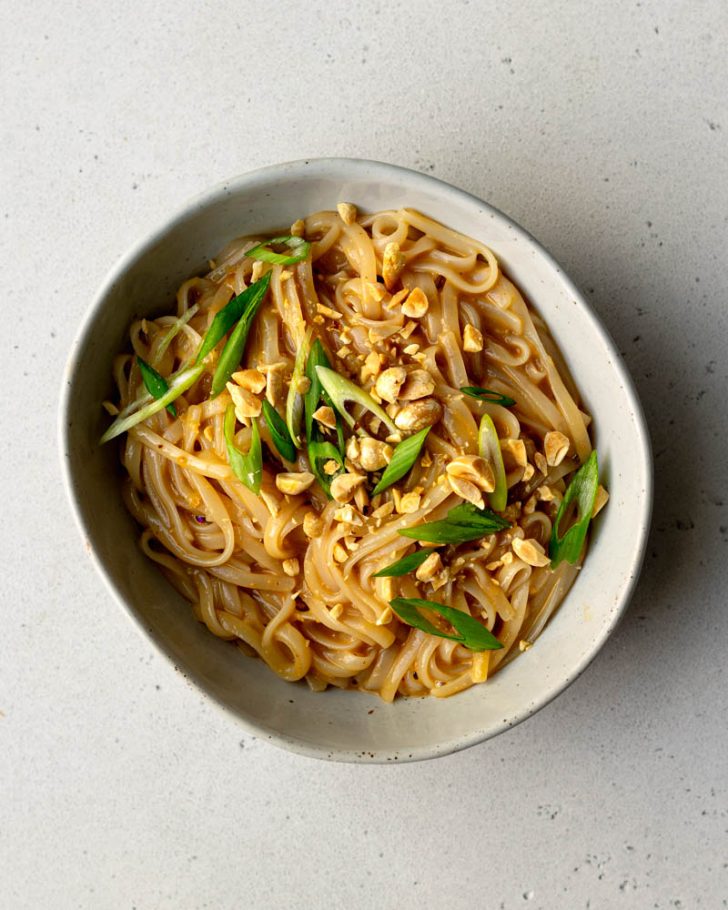 bowl of rice noodles with peanut sauce topped with scallions and crushed peanuts on a light grey backdrop