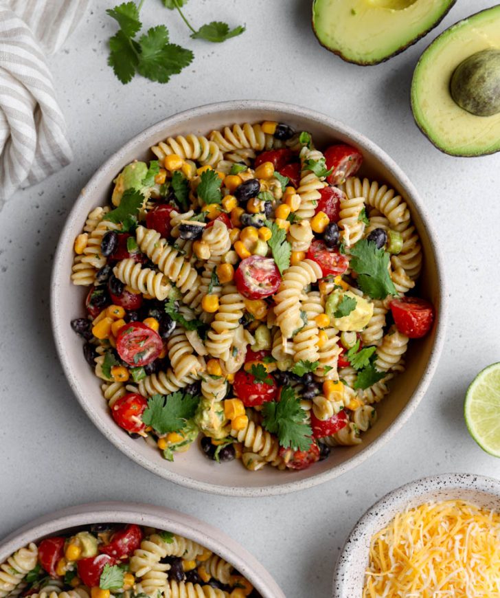 bowl of taco pasta salad on a light grey backdrop surrounded by ingredients like avocado, bowl of cheese, and cilantro leaves