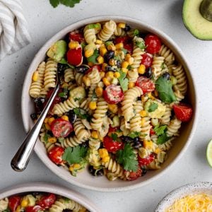 bowl of taco pasta salad with a silver fork on a light grey backdrop surrounded by ingredients like avocado, bowl of cheese, and cilantro leaves