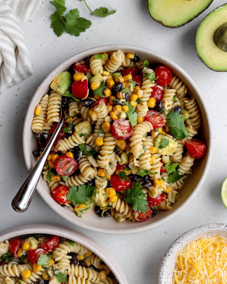 bowl of taco pasta salad with a silver fork on a light grey backdrop surrounded by ingredients like avocado, bowl of cheese, and cilantro leaves