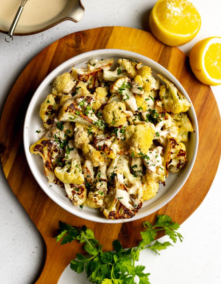 bowl of tahini cauliflower on a wooden cutting board surrounded by halved lemons, parsley, and a bowl of tahini sauce