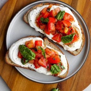 Two pieces of tomato basil whipped cottage cheese toast on a plate on a round wooden cutting board