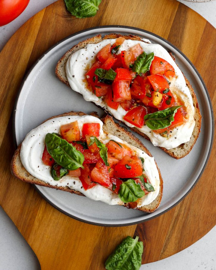Two pieces of tomato basil whipped cottage cheese toast on a plate on a round wooden cutting board