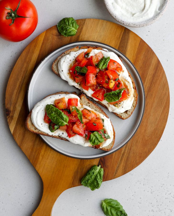 Two pieces of tomato basil whipped cottage cheese toast on a plate on a round wooden cutting board