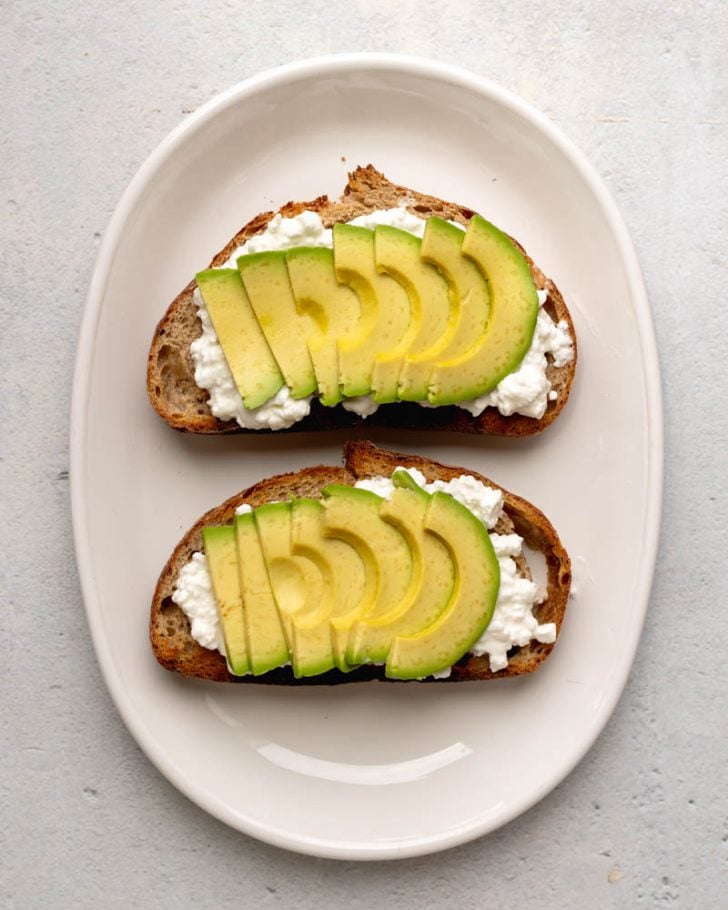 two pieces of toast with cottage cheese and avocado slices on a white oval plate